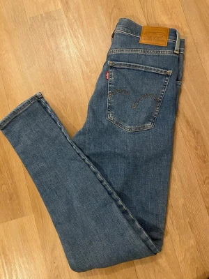 Levi's blå skinny jeans W26 L30 - Säljer ett par klassiska blå skinny jeans från Levi's med snygga detaljer på bakfickorna. Jeansen har normalhög midja och är tillverkade i bomull med lite stretch för extra komfort. Perfekta till sneakers eller boots.