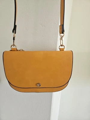 Gul axelväska med justerbart band - Snygg gul axelväska i slätt konstläder med rundad form och svart kantdetalj. Väskan har ett justerbart axelband och stängs med en knapp framtill. Perfekt för dig som vill ha en stilren och trendig accessoar till din outfit.
