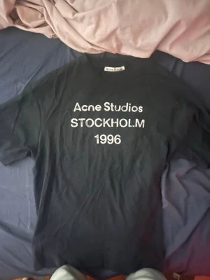 Svart Acne Studios t-shirt - Säljer en svart t-shirt från Acne Studios med vit text på bröstet där det står 'Acne Studios STOCKHOLM 1996'. Klassisk passform och rund halsringning. Perfekt för dig som gillar stilrena och ikoniska plagg. Kvitto finns.