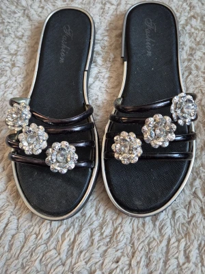 Svarta sandaler med blommiga strassdetaljer - Snygga svarta sandaler med tre tunna remmar och stora blommiga strassdetaljer på ovansidan. Sandalerna har en platt sula och silverfärgade kanter som ger en extra touch. Perfekta för att lyfta din sommarstil.