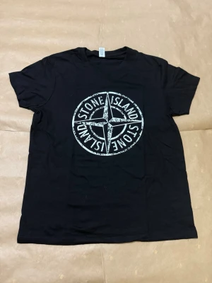 Svart Stone Island T-shirt - Svart t-shirt från Stone Island med stort vitt logotryck på bröstet. Klassisk rund hals och korta ärmar. Perfekt för dig som gillar streetwear och vill ha en stilren look. T-shirten är helt ny och har aldrig använts. Storlek S