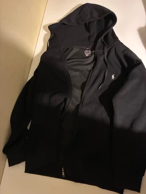 Svart hoodie från Polo Ralph Lauren - Säljer en svart hoodie från Polo Ralph Lauren med dragkedja framtill och klassisk huva. Tröjan har det ikoniska vita Polo-logotrycket på bröstet och är tillverkad i mjukt bomullsmaterial. Perfekt för en avslappnad och stilren look.