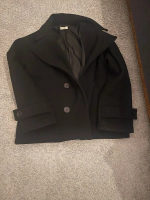 Svart kappa från Design by si - Elegant svart kappa från Design by Si med stilren siluett och bred krage. Modellen har knäppning framtill och dekorativa detaljer på ärmarna. Endast använd en gång och i mycket bra skick – som ny. Perfekt ytterplagg till våren☀️💐. 