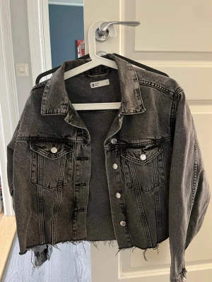 Svart croppad jeansjacka - Säljer en svart croppad jeansjacka från Gina Tricot med rå kant nertill och klassiska bröstfickor med knappar. Jackan har en cool, tvättad look och silverfärgade knappar framtill. Perfekt för dig som vill ha en edgy stil.