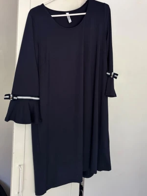 Marinblå klänning med volangärm - Mörkblå klänning med rund halsringning och långa ärmar. Ärmarna har volang och dekoreras med vita band och små rosetter. Enkel och stilren design som passar till många tillfällen. Storlek L. 