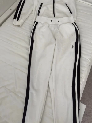 Vit tracksuit från Lion Athletics - Säljer en vit tracksuit från Lion Athletics med svarta ränder längs ärmar och ben. Setet består av ziptröja med fickor och matchande byxor med resår i midjan. Materialet är mjukt och bekvämt, perfekt för chill dagar hemma.