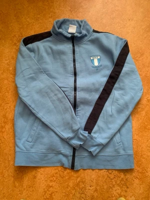 Ljusblå Malmö FF ziptröja - Ljusblå ziptröja från Malmö FF med klubbens logga broderad på bröstet. Tröjan har svarta detaljer längs axlar och ärmar, hög krage och två fickor framtill. Perfekt för dig som vill visa ditt stöd för laget.