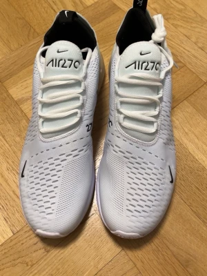 Nike Air Max 270 vita sneakers - Nike Air Max 270 sneakers i vitt med svart logga och detaljer. Skorna har en luftig mesh-överdel, synlig Air-enhet i hälen och snörning. Snygg och sportig design som passar perfekt till streetwear.
