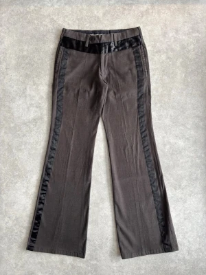 Tornado Mart kostymbyxor med satinränder - Snygga bruna kostymbyxor med svarta satinränder längs sidorna och satin i midjan. Byxorna har utsvängda ben och bälteshällor. Perfekta för dig som vill ha en unik twist på klassiska kostymbyxor.