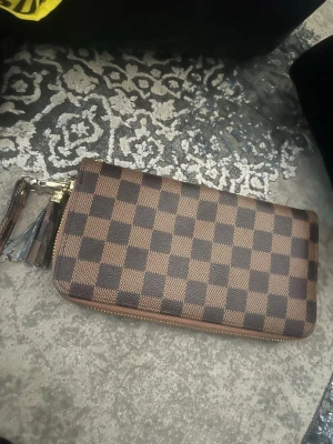 Brun plånbok med rutigt mönster - Snygg brun plånbok med klassiskt rutigt mönster i mörkbrunt och ljusbrunt. Plånboken har dragkedja i guldton och en matchande handledsrem med tofsdetalj. Tillverkad i konstläder med stilren design, perfekt för att hålla koll på kort och kontanter.