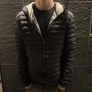 Svart pufferjacka från Colmar - Säljer en stilren svart pufferjacka från Colmar. Jackan har en dragkedja framtill och en huva för extra skydd. Perfekt för kyligare dagar med sin quiltade design som håller värmen. Märkeslogga på ärmen ger en snygg detalj. Galge medföljer