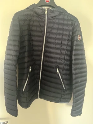 Marinblå Colmar dunjacka - Snygg marinblå dunjacka från Colmar med huva och två dragkedjeförsedda fickor framtill. Jackan har quiltad design och smala kanaler, samt en logotyp på ärmen. Perfekt för kalla vinterdagar och enkel att matcha med olika outfits. Jackan har inte använts och är i 10/10 skick, storlek 36. Passar för de som har storlek S. 
