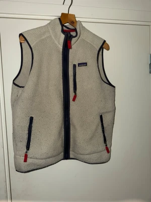 Beige fleeceväst från Patagonia - Mysig beige fleeceväst från Patagonia med marinblå detaljer och röda dragkedjor. Västen har hög krage, två fickor framtill och en bröstficka med dragkedja. Perfekt lager för kyliga dagar och enkel att matcha med olika outfits. Lite nopper på den men inget mer en det 😊