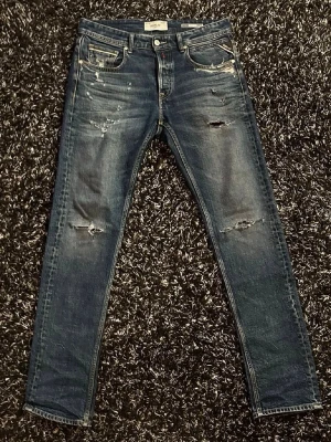 Blå slitna Replay jeans herr - Säljer ett par blå jeans från Replay med slitningar och distressed detaljer på både fram- och baksida. Klassisk femficksmodell med raka ben och läderpatch bak i midjan. Jeansen har en snygg tvättad look och är tillverkade i denim.