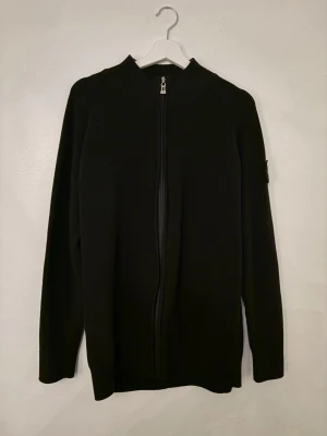 Svart ziptröja från Stone Island - Snygg svart ziptröja från Stone Island med hel dragkedja framtill och diskret logga på ärmen. Tröjan har ribbade detaljer och är tillverkad i ett mjukt, stretchigt material som passar perfekt till lager på lager. Enkel och stilren design.