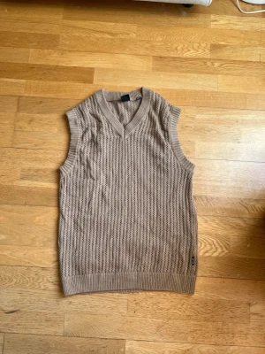 Beige stickad väst från Only & Sons - Snygg beige stickad väst från Only & Sons med v-ringad hals och ribbade kanter. Västen har ett diskret hålmönster och är perfekt för lager på lager-stil. Passar till både skjorta och t-shirt för en trendig look.