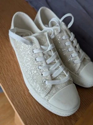 Glittriga vita låga sneakers stlk 37 - Säljer ett par vita sneakers med glitterdetaljer och vita snören. Skorna har en klassisk låg modell med rund tå och vit gummisula. Perfekta för skolavslutningen. Endast använda fåtal gånger. Stlk 37.