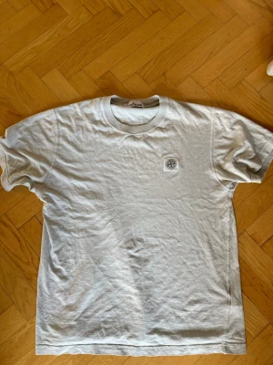 Ljusblå t-shirt från Stone Island - Ljusblå t-shirt från Stone Island med klassisk logopatch på bröstet. T-shirten har rund hals och korta ärmar, perfekt för en clean och stilren look. Tillverkad i mjuk bomull som känns skön mot huden. Säljer eftersom den är för liten