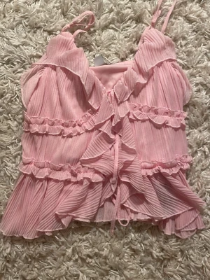 Rosa volangtop med smala axelband - Supersöt rosa topp med tunna axelband, volanger och plisserade detaljer. Toppen har v-ringning och knytning framtill samt flera lager volanger som ger en romantisk och lekfull känsla. Perfekt för dig som gillar en feminin och trendig stil. Aldrig använd! Nytt skick! Köp gärnaaa! Tröjan är i storlek S men passar för folk med Xs.