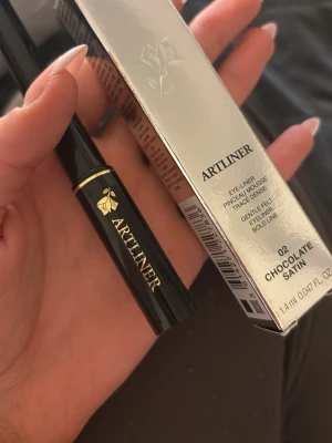 Lancôme Artliner 02 Chocolate Satin - Lancôme Artliner i nyansen 02 Chocolate Satin är en flytande eyeliner med filtspets för exakt applicering. Färgen är en djup chokladbrun med satinfinish. Snygg svart och guldig förpackning med elegant design. Innehåller 1,4 ml.