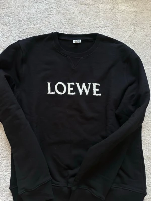 Svart sweatshirt från Loewe - Svart sweatshirt från Loewe med vit broderad logga framtill. Tröjan har rund halsringning och ribbade muddar vid ärmslut och nederkant. Tillverkad i mjukt bomullsmaterial som känns skönt mot huden. Perfekt för en avslappnad och trendig look.