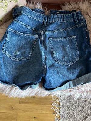 Blå high waist short för barn - Säljer ett par klassiska blå jeansshorts med upprullad kant och slitna detaljer. Shortsen har hög midja, fem fickor och stängs med dragkedja och knapp. Perfekta för varma dagar och en avslappnad look.