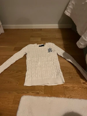Vit kabelstickad tröja från Ralph Lauren - Kabelstickad vit tröja från Ralph Lauren med rund halsringning och långa ärmar. Tröjan har ribbade muddar och nederkant samt en broderad RL-logga i mörkblått på bröstet. Perfekt för en klassisk och stilren look.