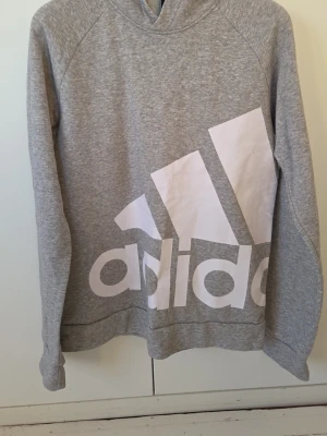 Grå hoodie från Adidas med stor logga - Snygg grå hoodie från Adidas med stor vit logga och text framtill. Hoodien har huva och är tillverkad i mjukt sweatshirtmaterial. Perfekt för en avslappnad och sportig stil. Långa ärmar och klassisk passform.