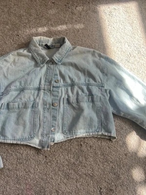 Ljusblå cropped jeansjacka - Trendig ljusblå cropped jeansjacka med klassisk krage och stora framfickor. Jackan har knappstängning framtill och raka ärmar. Perfekt för dig som gillar en avslappnad och modern stil.