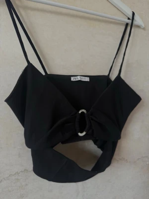 Svart croppad topp från Zara - Trendig svart croppad topp från Zara med smala axelband och en snygg cut-out framtill. Toppen har en dekorativ ringdetalj i mitten och är tillverkad i ett mjukt material som sitter bekvämt. Perfekt för dig som vill sticka ut med en edgy look.