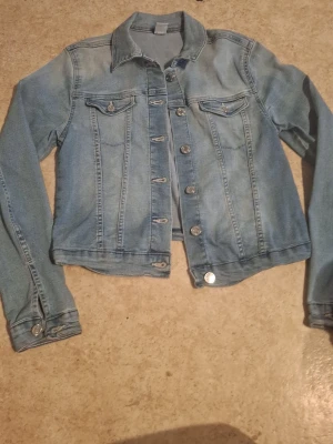 Ljusblå klassisk jeansjacka - Snygg ljusblå jeansjacka med raka sömmar, två bröstfickor med lock och silverfärgade knappar framtill. Jackan har en klassisk krage och långa ärmar med knapp vid ärmslut. Perfekt att slänga över en hoodie eller t-shirt.