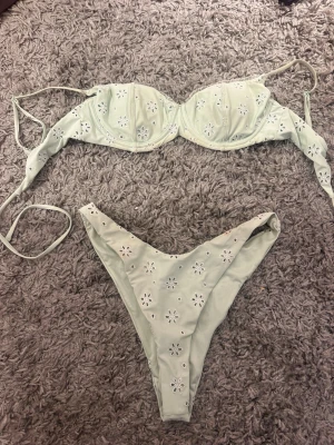 Ljusgrön bikini med blommiga detaljer - Söt ljusgrön bikini med broderade blommor och håldetaljer. Överdelen har bygel och knyt i ryggen, medan underdelen har en klassisk passform. Perfekt för sommaren och strandhäng!