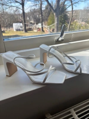 Klackar  - Säljer ett par stilrena vita sandaletter med fyrkantig tå och tunna remmar. Skorna har en hög blockklack och öppen häl, vilket ger en modern och trendig look. Perfekta för dig som vill ha en clean och minimalistisk stil. Materialet ser ut att vara konstläder.
