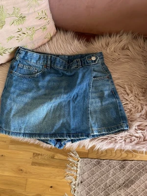 Blå jeansskort med fickor - Snygg blå jeansskort i klassisk denim med framfickor och bakfickor. Skorten har en omlott-look framtill, bälteshällor och stängs med knapp och dragkedja. Perfekt för en avslappnad och trendig stil.