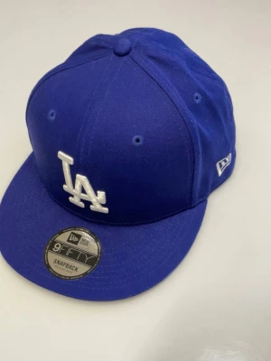 Blå LA Dodgers keps från New Era - Snygg blå snapback-keps från New Era med LA Dodgers-logga broderad i vitt framtill. Modellen är 9FIFTY och har en rak skärm samt justerbar baksida. Perfekt för dig som vill ha en klassisk och sportig look. 53 cm runt i kepsen och kan förstoras.