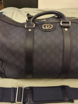 Gucci svart weekendbag med logga - Snygg svart weekendbag från Gucci med GG-monogram, dubbla topphandtag och avtagbar axelrem. Väskan har detaljer i läder, dragkedja upptill och två innerfack. Fodrad med bomullslinne och har metallogo framtill. Perfekt för resor och har plats för allt du behöver. Orginal-hänglås medföljer.