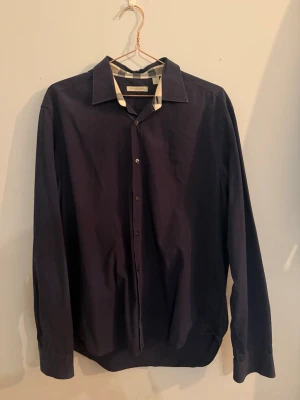 Burberry Brit skjorta – mörkblå – strl XL- passar L - Elegant skjorta från Burberry Brit i en mörkblå nyans med klassiska rutiga detaljer i kragen samt vid handlederna. En tidlös modell med diskret broderad logotyp på bröstet.  Storlek: L Färg: Mörkblå Skick: 8/10  📦 Kan skickas 💬 Kontakta vid frågor eller intresse