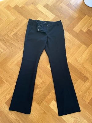 Svarta lågmidjade kostymbyxor - Snygga svarta lågmidjade kostymbyxor med pressveck. Byxorna har hällor för skärp, dold knäppning fram och två fuskfickor bak. Perfekta för en clean och stilren look.