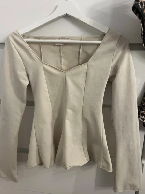 Beige långärmad topp Gina Tricot - Säljer en beige långärmad topp från Gina Tricot med v-ringad hals och figurnära passform. Toppen har en lätt utsvängd nederdel och sömdetaljer framtill som ger en snygg siluett. Perfekt att styla med jeans eller kjol.