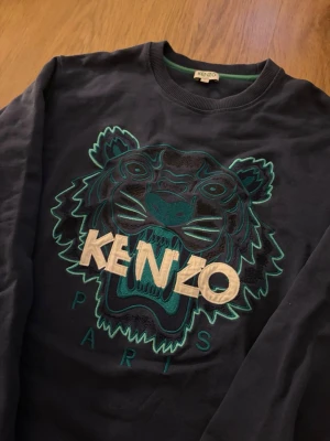 Kenzo Tröja - Snygg mörkblå sweatshirt från Kenzo med det ikoniska tigerhuvudet broderat i grönt, svart och vitt på bröstet. Rund halsringning och långa ärmar. Stort KENZO-tryck och PARIS-broderi under motivet. Perfekt för dig som gillar streetwear och statement-plagg. Storleken är S men passar även XS. Tröjan är fint skick.                                             Skriv om ni har några funderingar!😄