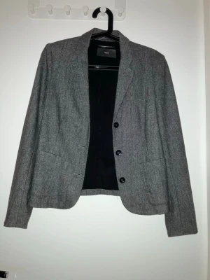 Grå kavaj från Filippa K - Stilren grå kavaj från Filippa K med klassisk krage, tre svarta knappar och två stora framfickor.