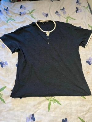 Mörkblå t-shirt med vita detaljer - Snygg mörkblå t-shirt från Ecko Unltd. med vita kontrastdetaljer vid halsringning och ärmslut. T-shirten har korta ärmar och knappar vid halsen för en avslappnad look. Perfekt till jeans eller shorts.