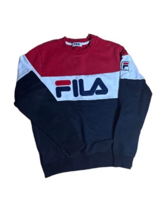 FILA färgblockad stickad tröja - Snygg stickad tröja från FILA i färgblockad design med rött, vitt och marinblått. Stor FILA-logga framtill och mindre logga på ärmen. Tröjan har rund halsringning och långa ärmar, perfekt för en sportig och trendig look.