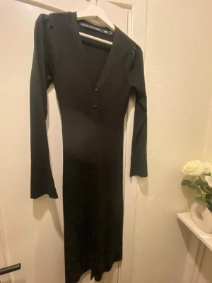 Svart ribbad långklänning med knappar - Svart långklänning i ribbad stickad kvalitet med v-ringning och knappar framtill. Klänningen har långa ärmar och en smickrande slim passform. Perfekt för dig som gillar stilrena och tidlösa plagg.