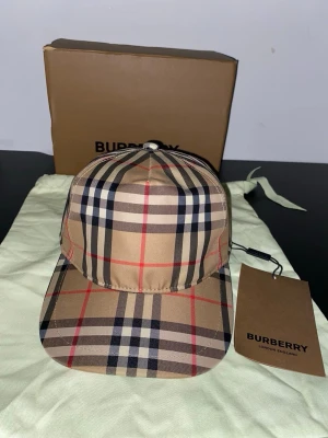 Burberry keps - Säljer en snygg keps från Burberry i deras ikoniska beigea rutmönster med svarta, vita och röda linjer. Kepsen har böjd skärm och justerbar rem bak. Tillverkad i ett slätt tyg som känns lyxigt och stilrent. Perfekt accessoar för att lyfta din outfit.