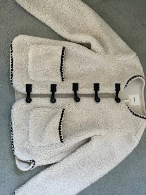 Krämvit teddyjacka från .OBJECT - Säljer en krämvit teddyjacka från .OBJECT med stora framfickor och svarta dekorativa kanter. Jackan har rund halsringning och stängs med svarta knappar i toggle-stil. Mysigt, fluffigt material som ger en trendig look.