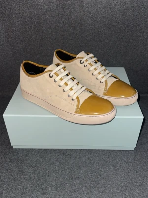Lanvin beige sneakers - Snygga sneakers från Lanvin i beige mocka med glansig tå och matchande detaljer. Skorna har vita snören, lågt skaft och klassisk gummisula med Lanvin-logga undertill. Perfekta för dig som gillar stilrena men unika sneakers.