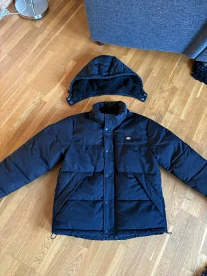Svart dunjacka från Dickies - Säljer en svart dunjacka från Dickies med avtagbar huva. Jackan har hög krage, tryckknappar och dragkedja framtill samt två stora fickor. Perfekt för kalla vinterdagar och riktigt snygg streetstil.