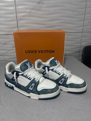 Louis Vuitton Trainers  - Snygga Louis Vuitton sneakers i vitt och mörkblått med klassiskt LV-mönster på sidorna. Skorna har tydliga logodetaljer, snörning och en chunky sula. Materialmix av skinn och textil ger en exklusiv känsla. Perfekta för dig som vill sticka ut med lyxig streetstyle.