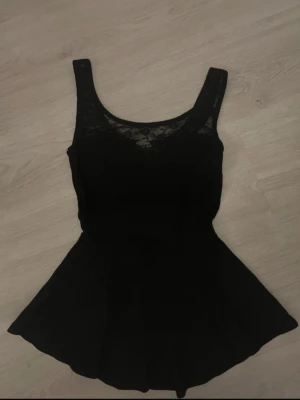 Svart top med spetsdetalj - Snygg svart peplumtop med breda axelband och v-ringad spets upptill. Toppen har en figurnära passform upptill och utställd nederdel som ger en feminin siluett. Superfin!